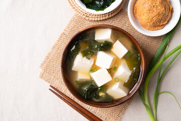 Top view of healthy Japanese miso soup with soft tofu and seaweed in a wooden bowl with chopsticks on white fabric background