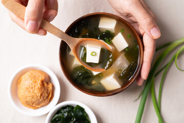 Top view of hand holding spoon and eating healthy Japanese miso soup with soft tofu and seaweed in a wooden bowl on white fabric background