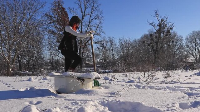A woman in winter clothes stands in a snowy bathtub in the yard, leaning on a shovel, looking into the distance on a frosty sunny day.