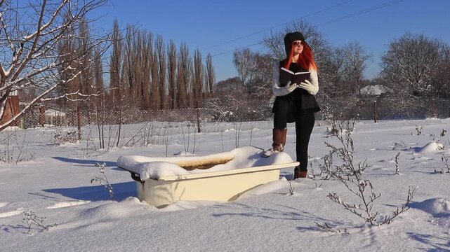 A beautiful girl with a diary in her hands near an old bathtub in the middle of a snowy landscape plans her day. A combination of fashion, nature and an old bathtub.