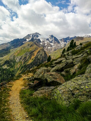 Obraz premium Vertical View of a Narrow Alpine Trail Along a Rocky Ridge with Snowy Peaks, Dramatic Clouds and Tree Line in the High Mountains of Trentino Alto Adige, Italy