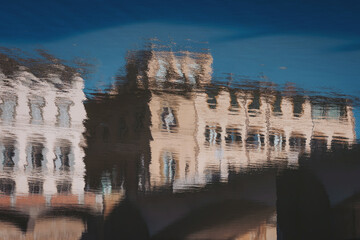 Buildings are reflected in water, showing distorted shapes and colors. The scene takes place in a city during daylight, with a clear blue sky above