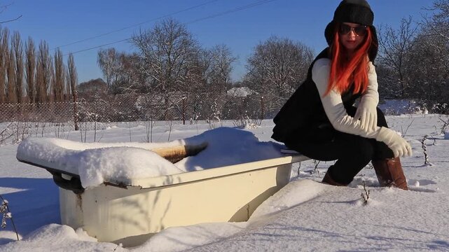 Stylish woman with red hair poses near an old bathtub in a snowy landscape. A combination of fashion, nature and an old bathtub. Winter fashion mood, contrast of style and cold.