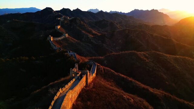 Aerial view of the majestic Great Wall snaking through the rugged, mountainous terrain, bathed in the warm glow of the setting sun, Huairou District, China.