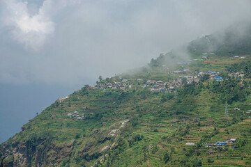 Fototapeta premium AERIAL: White clouds drifts over the rural village on the steep hill in Nepal.