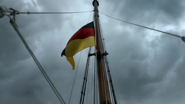 Gusting wind lifting German tricolor flag, unfurling and whipping along mast and rigging at sea
