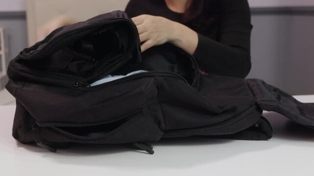 Female hands placing folded clothes into black backpack on white table indoors, close up view symbolizing travel packing, organization and preparation for trip.