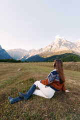 Obraz premium woman in sweater and boots sitting in meadow, mountains landscape and nature around her: young female with longhair relaxes on grassy field at dusk, enjoying serene outdoor solitude and scenic view