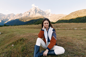 Obraz premium Woman portrait sitting in meadow with mountains in background, wearing shearling jacket and rubber boots, autumn outdoor scene, peaceful nature, relaxed young traveler.