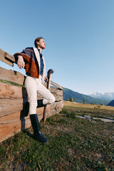 Obraz premium Woman leaning on wooden fence in countryside mountains, portrait outdoors wearing jacket and tall boots, looking into the distance during travel and nature lifestyle.