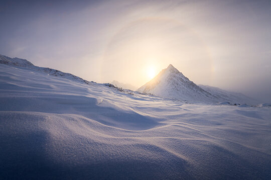 View of a snow-blanketed landscape under a haloed sun, where crisp, undulating snowdrifts meet a majestic, pyramid-shaped mountain peak, Coldfoot, Alaska, United States.