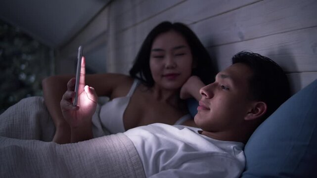 Couple in bed using smartphone, night bedroom light. Addiction social media digital technology, relationship coldness issue. Cell mobile phone scrolling problem. Useless cellphone internet surfing.