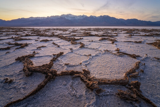 View of cracked earth stretches into the horizon under a pastel sky, with snow-capped mountains in the distance, Stovepipe Wells, CA, United States.