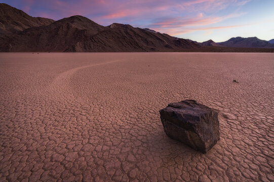 View of a solitary rock resting on the cracked, dry lakebed under a pastel sunset sky, Stovepipe Wells, CA, United States.