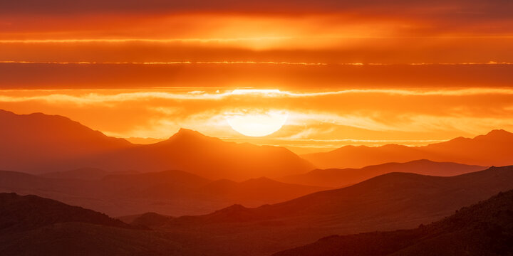 View of a fiery sunset bathes the silhouette of rolling mountains in a warm, orange glow, contrasting the textured sky in Stovepipe Wells, CA, United States.