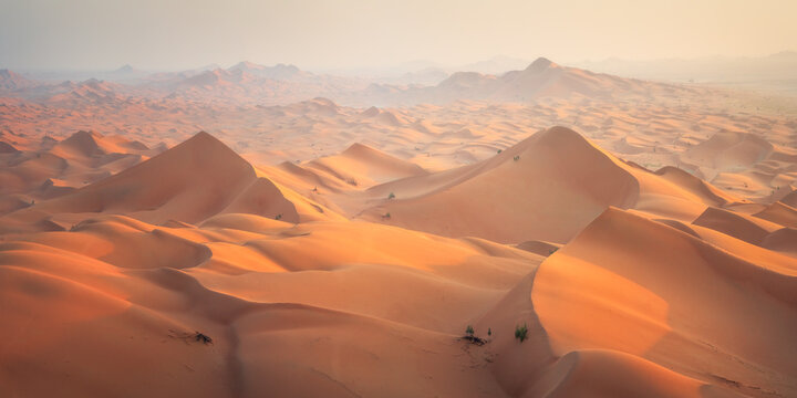 View of undulating dunes cast long shadows under a hazy sky, the warm hues of sand contrasting with the distant, muted mountains, Ubar, Dhofar Governorate, Oman.