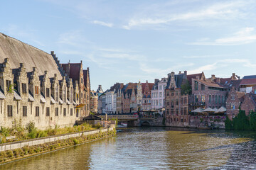 Fototapeta premium Buildings of varying shapes and colors line the riverbank in Ghent, Belgium. The large brick building is on the left. A bridge over the river is visible