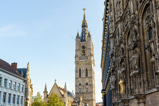 An ancient bell tower Belfried in the city of Ghent in Belgium The tower is made of stone with a black pointed roof and black tiles Round gold watch the facade of the building with Gothic patterns