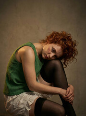 Artistic portrait of a melancholic ginger woman with curly hair, resting her head on her knee in a studio.
