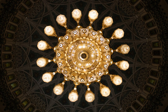 View of a grand chandelier with intricate patterns, casting a warm glow against the detailed ceiling, Muscat, Oman.