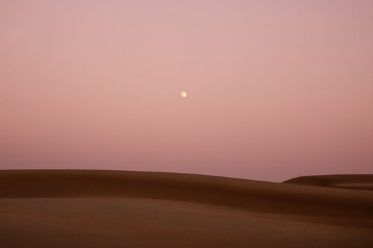 View of the warm, earthy dunes under a pale pink sky with a distant moon evoking a sense of serene isolation, Bidiyah, Ash Sharqiyah North Governorate, Oman.