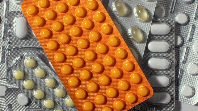 Rotating macro view of assorted pills in blister packs, orange tablets and capsules scattered on metallic foil background.