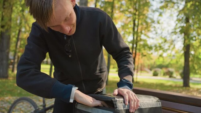 caucasian delivery rider inspecting bag, checking zipper and straps on park bench, bicycle parked nearby, autumn light filtering through trees, focused expression while preparing order for takeaway,