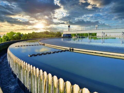 A large circular clarifier at a water treatment plant during sunset, with calm reflective water, overflow weirs, and elevated walkways surrounded by greenery and dramatic clouds.