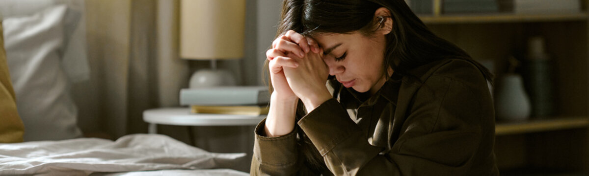 Website banner of young adult woman sitting on bed clasping hands together and bowing head with closed eyes, appearing deep in thought or prayer