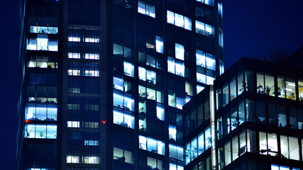 Obraz premium Big glowing windows in modern office building at night, in rows of windows light shines. Fragment of the glass facade of a modern corporate building at night. Modern glass office in city.