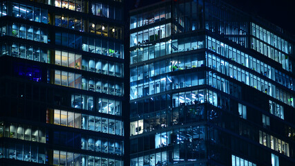 Fototapeta premium Big glowing windows in modern office building at night, in rows of windows light shines. Fragment of the glass facade of a modern corporate building at night. Modern glass office in city.
