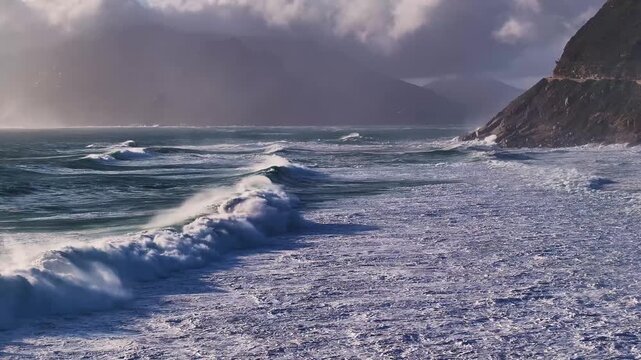 Aerial view of the coastline with waves crashing onto the shore under a cloudy sky, creating a dynamic interplay of light and shadow, Cape Town, Western Cape, South Africa.