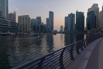 Naklejka premium Evening view of a river canal and a ship. Orange sunlight, skyscrapers, buildings and the embankment. On the streets of the Emirates, in a public place.
