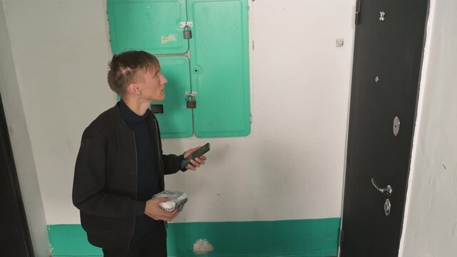young man delivering food at apartment door holding bag and checking phone, pressing buzzer and inspecting entry panel in narrow hallway, brief pause while locating apartment number, focused efficient