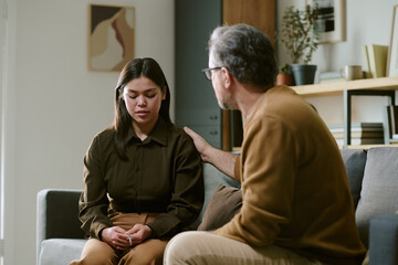 Young woman sitting on couch looking down while middle aged Caucasian pastor sitting beside her...