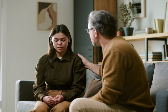 Young woman sitting on couch looking down while middle aged Caucasian pastor sitting beside her placing hand on her shoulder offering support in living room setting