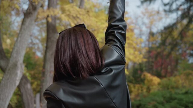 woman in leather jacket facing trees, reaching toward canopy with confident gesture back portrait shows textured hair and studded black jacket against yellow autumn leaves, park grove atmosphere,