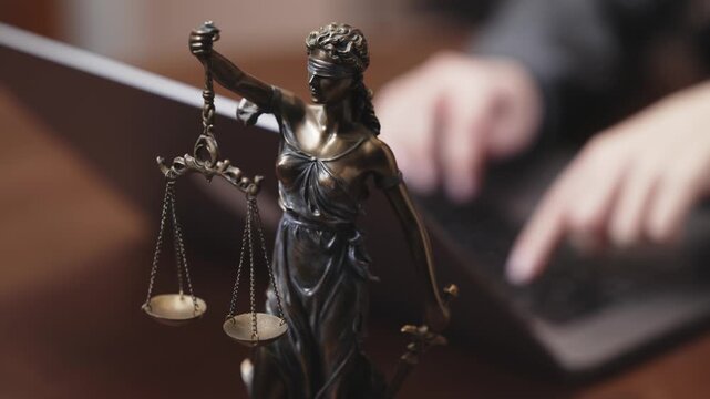 Bronze statue of Lady Justice holding scales in front of a laptop, symbolizing law and order in a modern legal setting with blurred hands typing in the background
