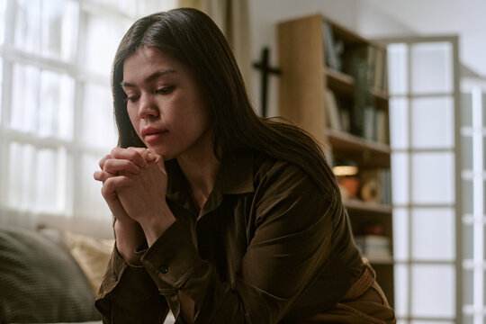 Young adult Asian woman sitting indoors clasping hands together with eyes closed appearing deep in thought or reflection, bookshelf and window in background, natural light illuminating scene