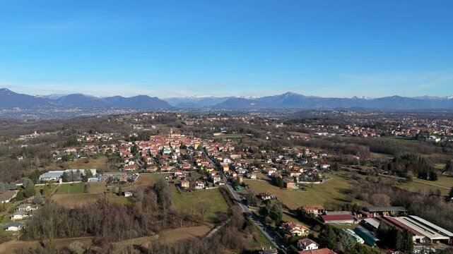 Aerial ascending view over the Varese area shows scattered towns, open countryside, and rolling terrain, with the Swiss Alps stretching across the horizon under clear skies.