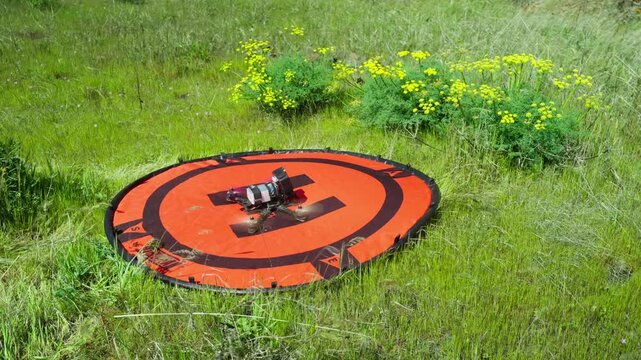 A First Person View (FPV) drone with a mounted GoPro action camera slowly taking off like a rocket from an orange hoodman helipad as the wind blows through green grass and yellow flowers