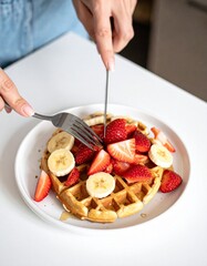 Primer plano de waffles reci&eacute;n hechos con banana y fresas, cortados con cuchillo y tenedor sobre un plato, luz natural y estilo culinario moderno.