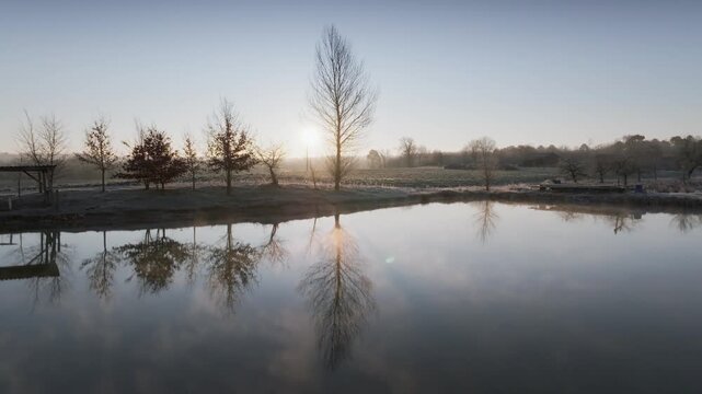 Low altitude drone shot gliding over still water reflecting bare trees at sunrise in Monpazier France. Winter landscape with morning mist and golden light illuminating rural nature scene.