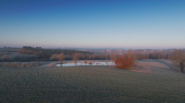 High angle drone view of frosty fields and small lake during dawn in French countryside. Golden hour light hitting bare trees surrounding water body in Monpazier winter landscape.