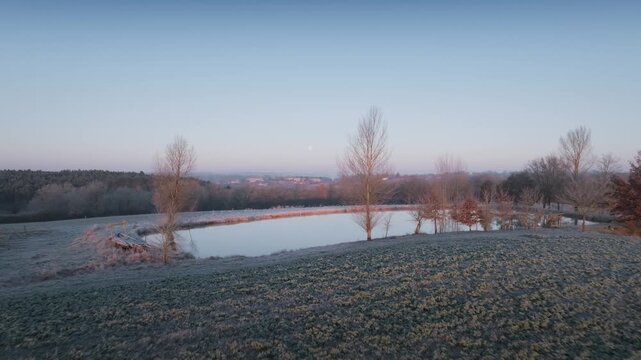 Low altitude drone shot gliding over still water reflecting bare trees at sunrise in Monpazier France. Winter landscape with morning mist and golden light illuminating rural nature scene.