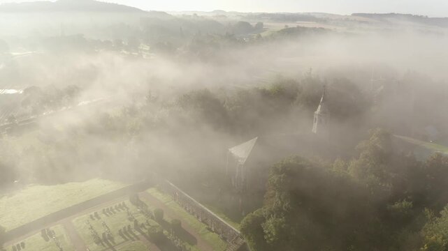 Aerial view of a church shrouded in a thick fog, its spire barely visible amidst the green trees, Cupar, Scotland, United Kingdom.