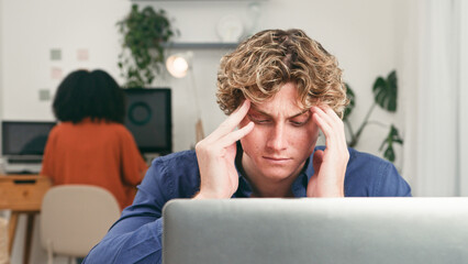Young Man Stressed at Laptop in Open Office With Startup Team Working Behind Him