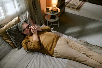 Obraz premium Middle aged Caucasian man lying on bed clasping hands together near face appearing thoughtful, wearing eyeglasses, relaxing in bedroom with soft lighting and cozy decor visible