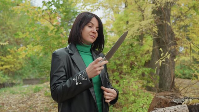 Leather jacket woman inspects knife outdoors among autumn trees by stacked logs and a bag, unfolding blade and studying edge with focused expression moody, solitary atmosphere combines rugged style