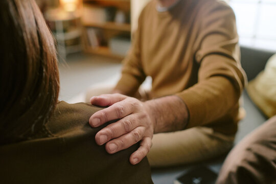 Middle aged man comforting middle aged Caucasian woman by placing hand on her shoulder, both sitting indoors, showing support and empathy during conversation